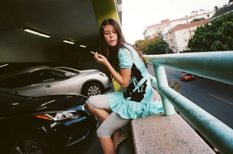 Young woman in blue dress sits on a concrete ledge near parked cars, holding a cigarette, with buildings and trees visible in the background.