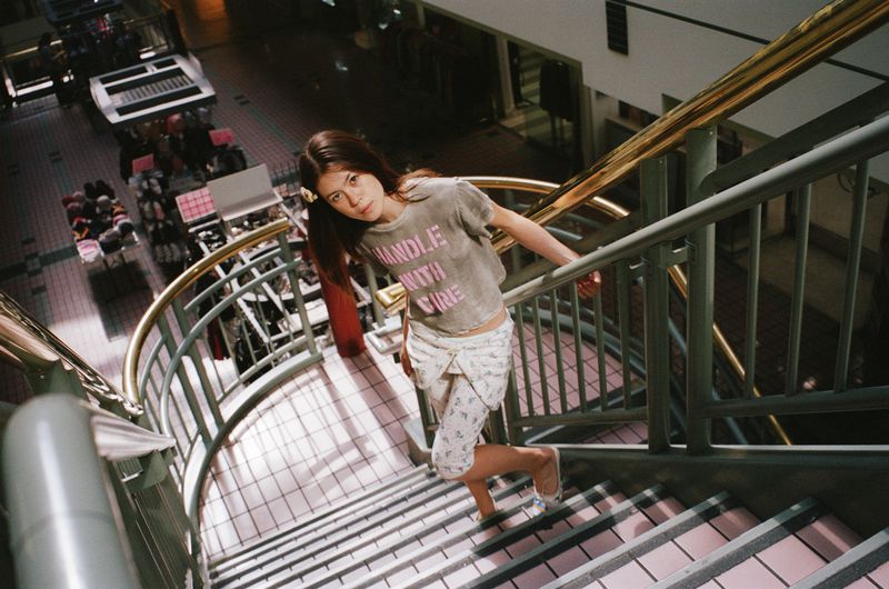 A young woman with long brown hair descends a staircase in an indoor shopping center wearing a t-shirt and patterned capri pants.