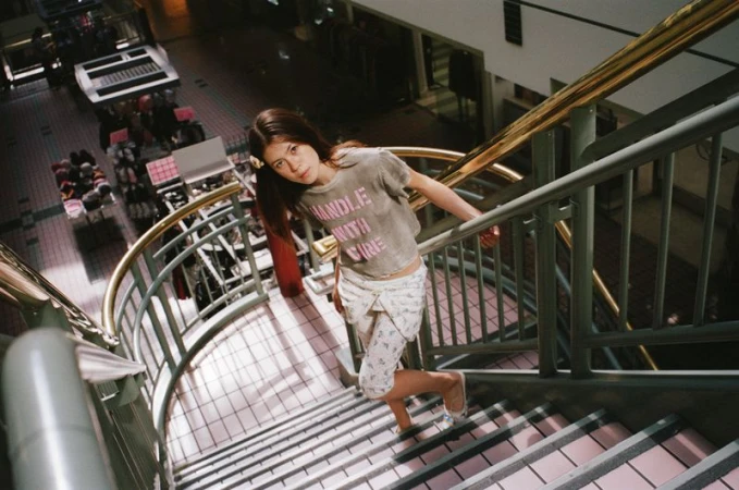 A young woman with long brown hair descends a staircase in an indoor shopping center wearing a t-shirt and patterned capri pants.