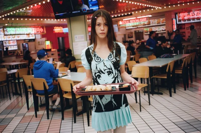 Young woman holding tray of food in a bustling food court. She is wearing a light blue dress with a distinctive cartoon design.