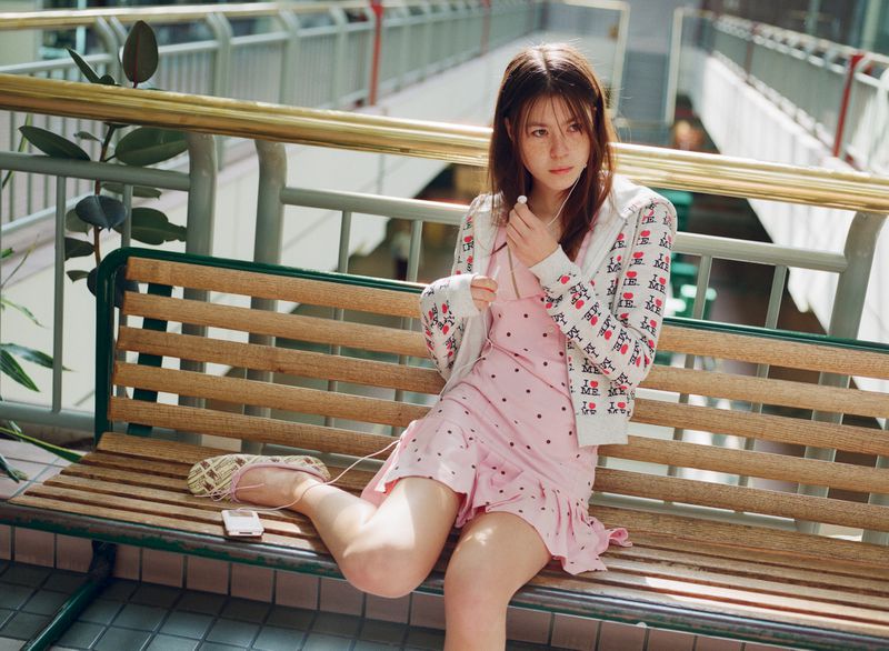 Young woman with earbuds in patterned jacket and polka-dot dress sits on a bench with phone in a public area with metal railings.