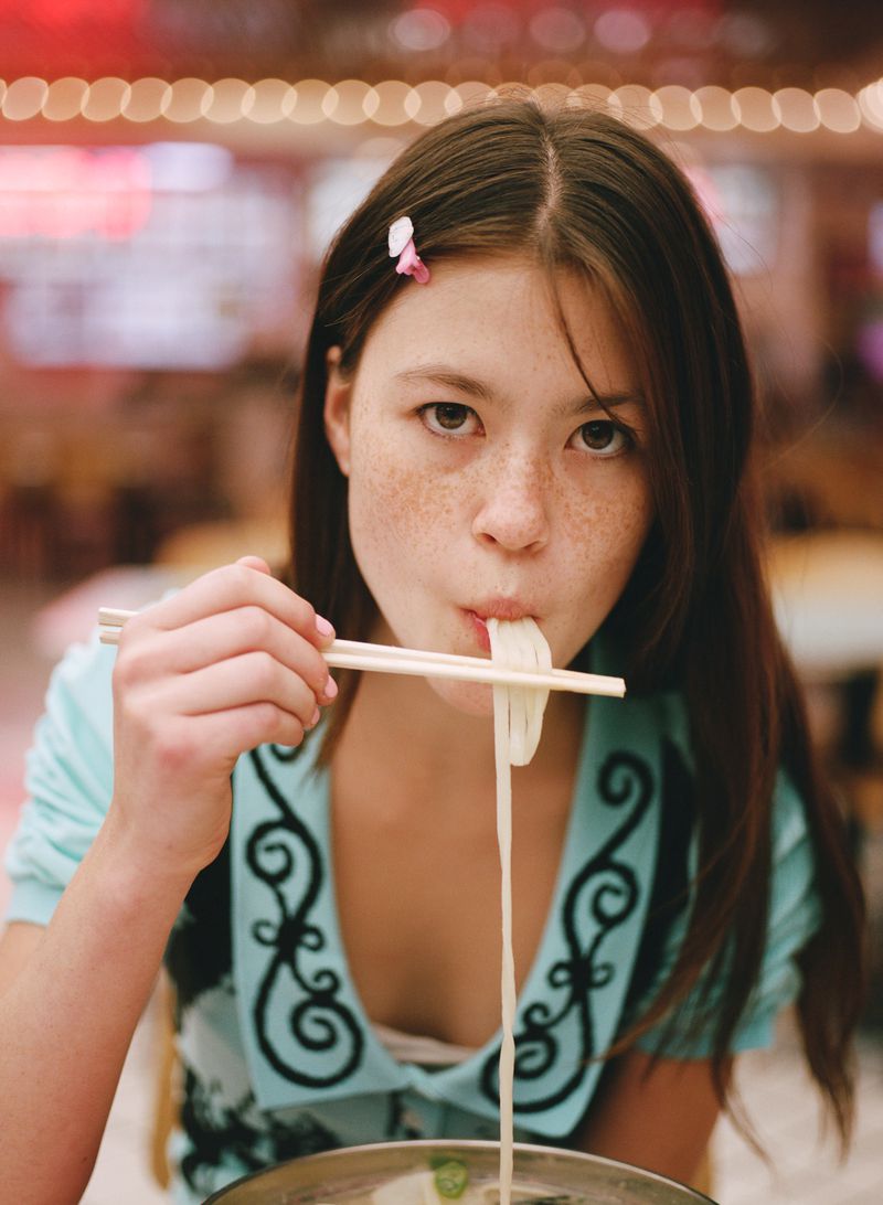 Girl with freckles enjoys noodles indoors, using chopsticks; wearing a light blue top with decorative designs. Bokeh lighting in background.