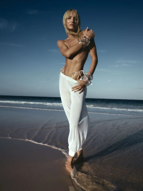 Adult woman poses in white pants and bracelets on a beach under a bright blue sky, standing in the shallow water with wet hair.