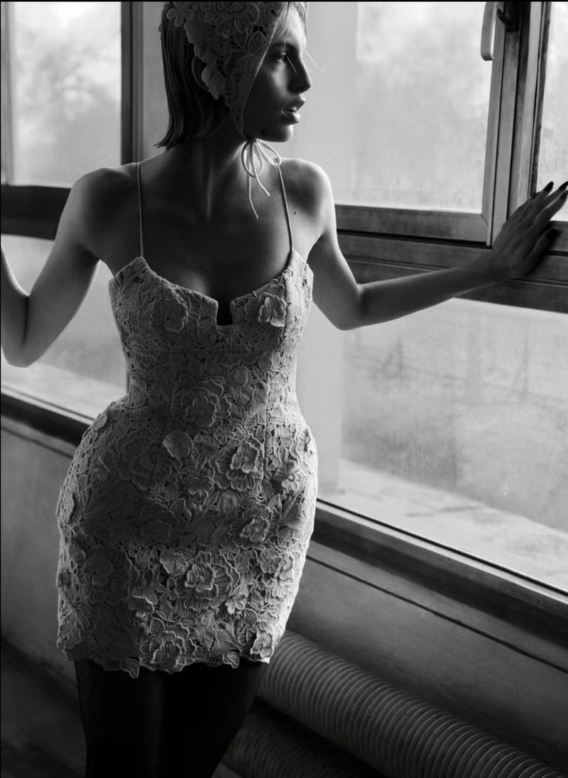 Woman in lace dress and bonnet looking out window in black and white. Indoor fashion portrait with natural light and window.