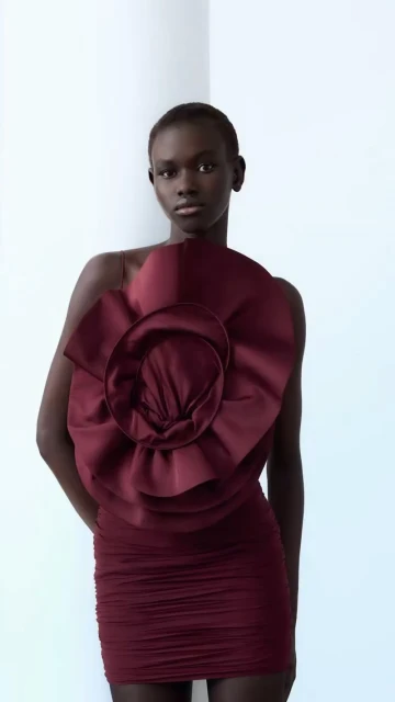 Elegant woman with dark skin poses in studio wearing a maroon dress with a large floral accent, complemented by a clean white backdrop.