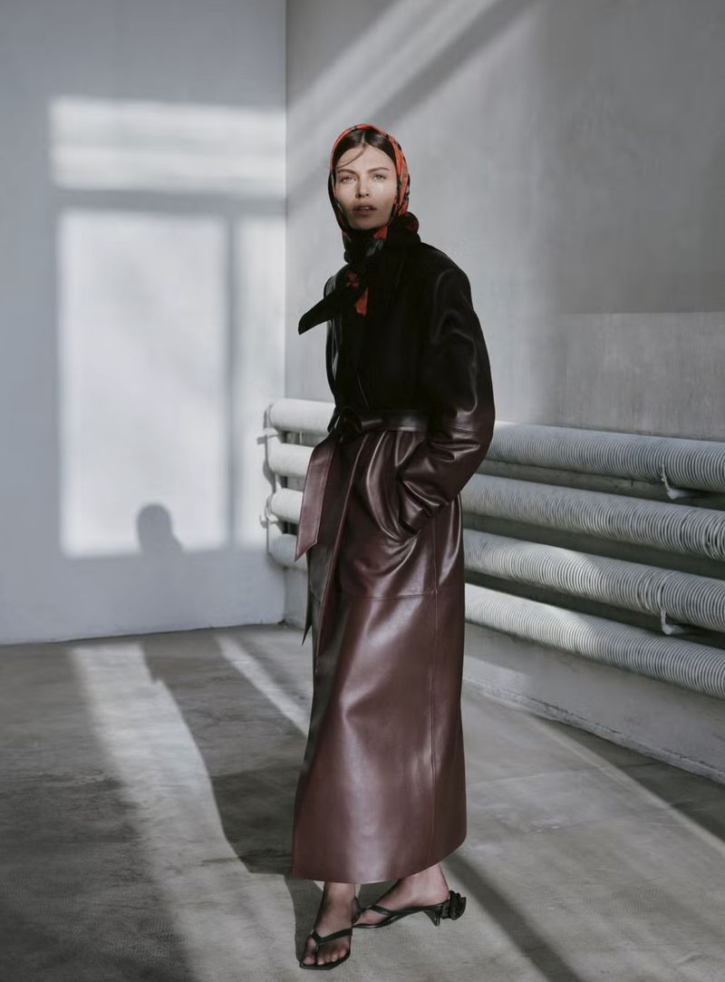 A woman in a brown leather coat and patterned headscarf stands in a corner of a room with light casting shadows on the wall and floor.