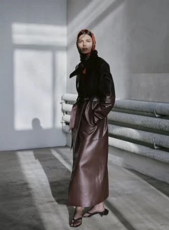 A woman in a brown leather coat and patterned headscarf stands in a corner of a room with light casting shadows on the wall and floor.