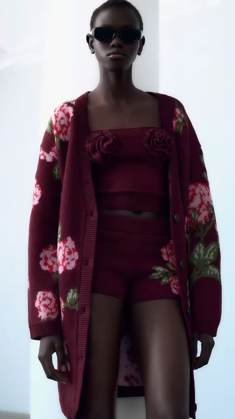 Woman poses in a matching burgundy knitwear set featuring floral embroidery in a studio with soft lighting and a blurred background.