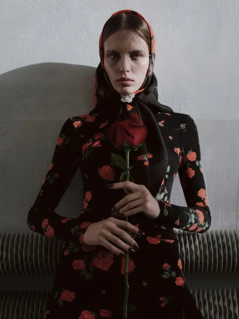 Elegant woman in floral dress and headscarf holding red rose. Indoor studio shot. Dark hair, pale skin, and moody lighting.