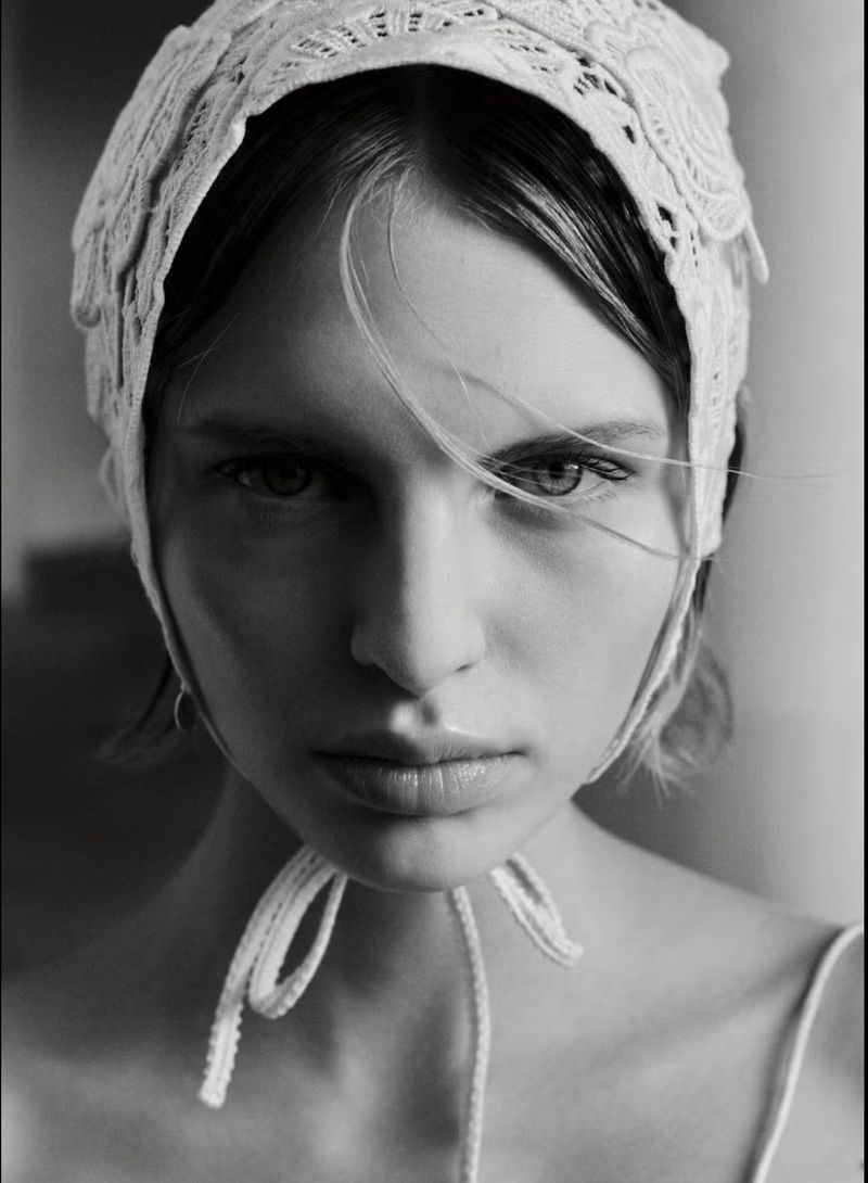 Striking black and white portrait of a woman wearing a lace bonnet, emphasizing her intense gaze and minimalist fashion style.