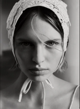 Close-up portrait of a model wearing a white lace bonnet with tied ribbons under the chin against an out-of-focus background.