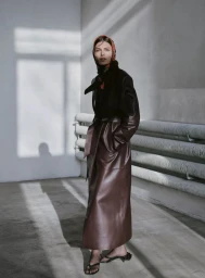 Model wearing a long leather coat and patterned headscarf standing in an interior space with natural light.