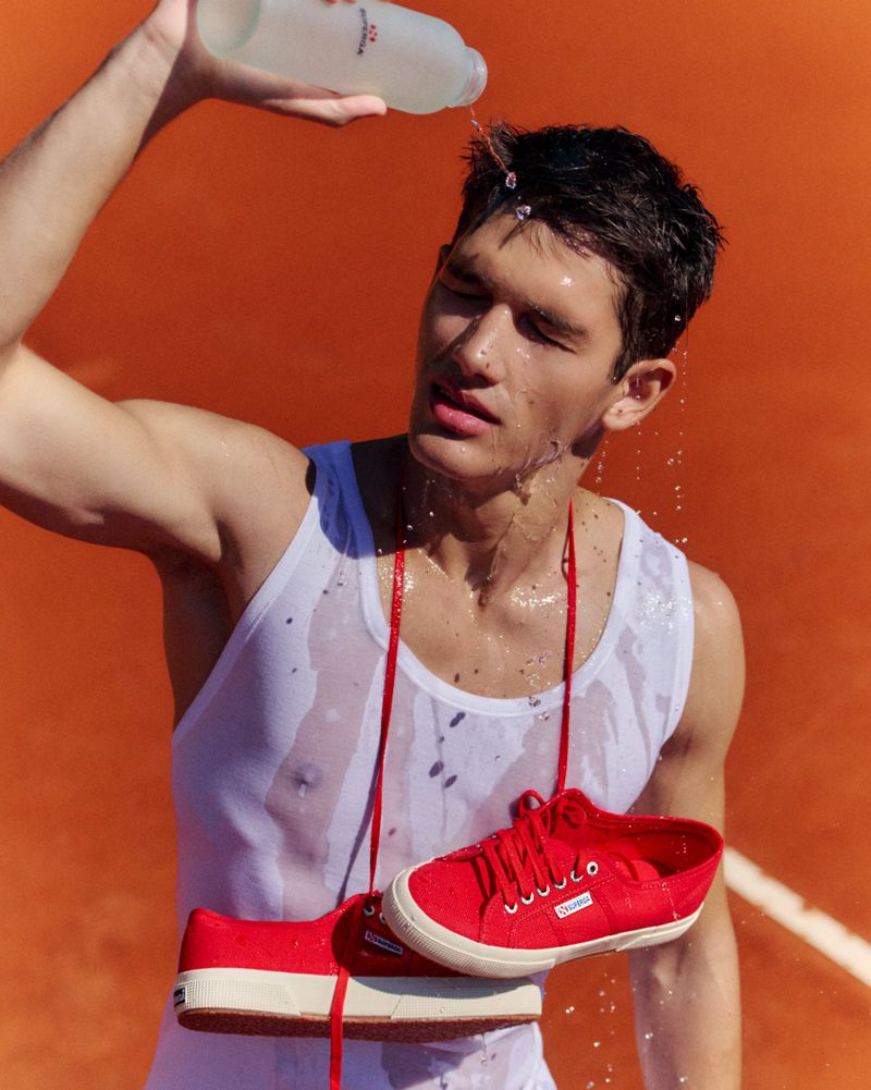 Athletic man in tank top is doused with water, refreshing after tennis. Red sneakers are slung around his neck. Bright, summery hues.