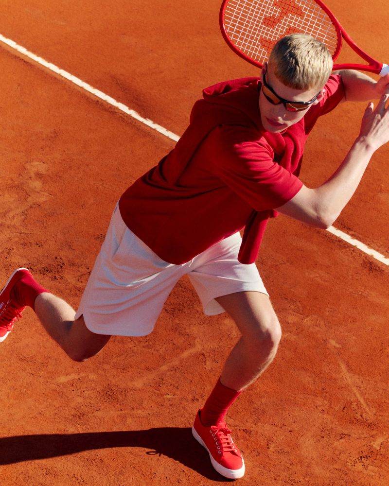 Man wearing red and white sportswear swinging a tennis racket on a clay court outdoors in bright sunlight.