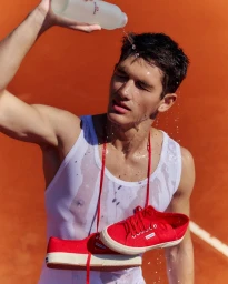 Man in white tank top pouring water on his head with red sneakers hanging around his neck against a clay court