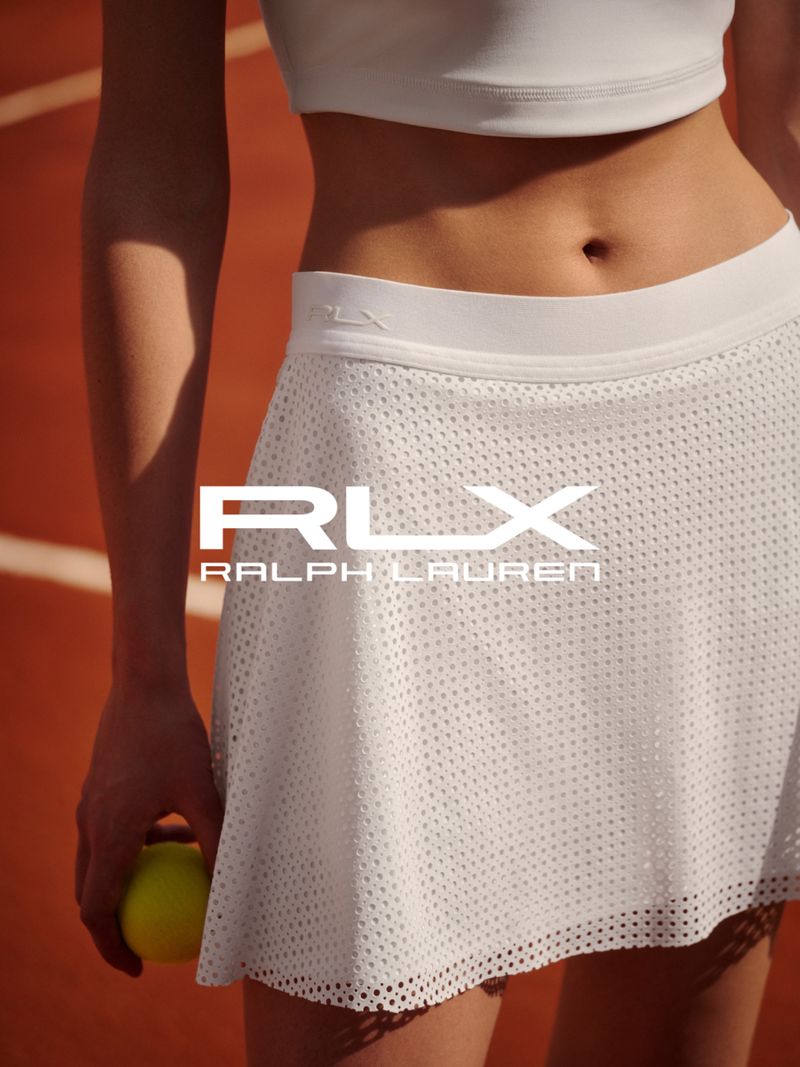 Athletic woman in white tennis outfit with RLX Ralph Lauren branding, holding a tennis ball, posing on a clay tennis court.