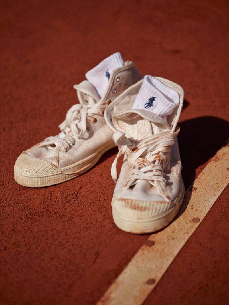 Close-up of white canvas sneakers with white Ralph Lauren socks, placed on a textured reddish-brown surface with a white line detail.