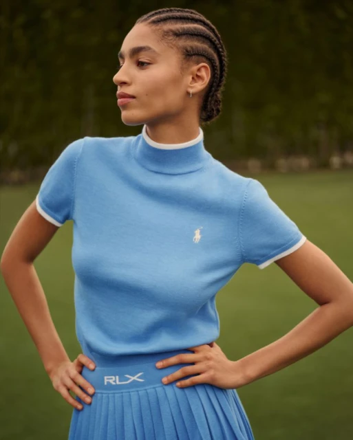 A woman poses in a light blue short-sleeve turtleneck and matching pleated skirt with her hands on her hips outside on grass.