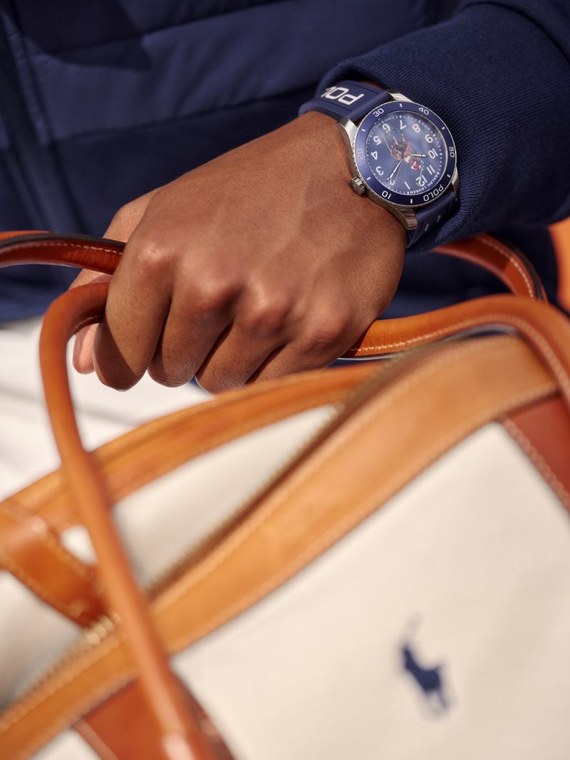Close-up shot of an adult's hand holding a bag with a watch visible. The watch has a blue face and strap and the bag is white and brown.