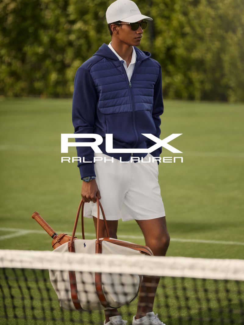A fashionable male model on a tennis court wearing athletic apparel and carrying a tennis bag, advertising Ralph Lauren's RLX line.