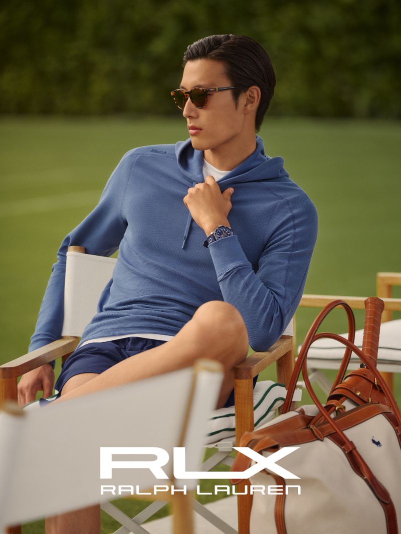 Asian male model in blue hoodie and sunglasses sits in a chair on a tennis court with Ralph Lauren RLX branded tennis bag.