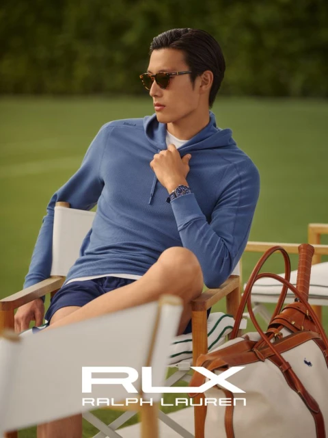 Asian male model in blue hoodie and sunglasses sits in a chair on a tennis court with Ralph Lauren RLX branded tennis bag.