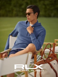 A man wearing a blue hoodie and sunglasses sits in a director chair outdoors with a tennis bag beside him.