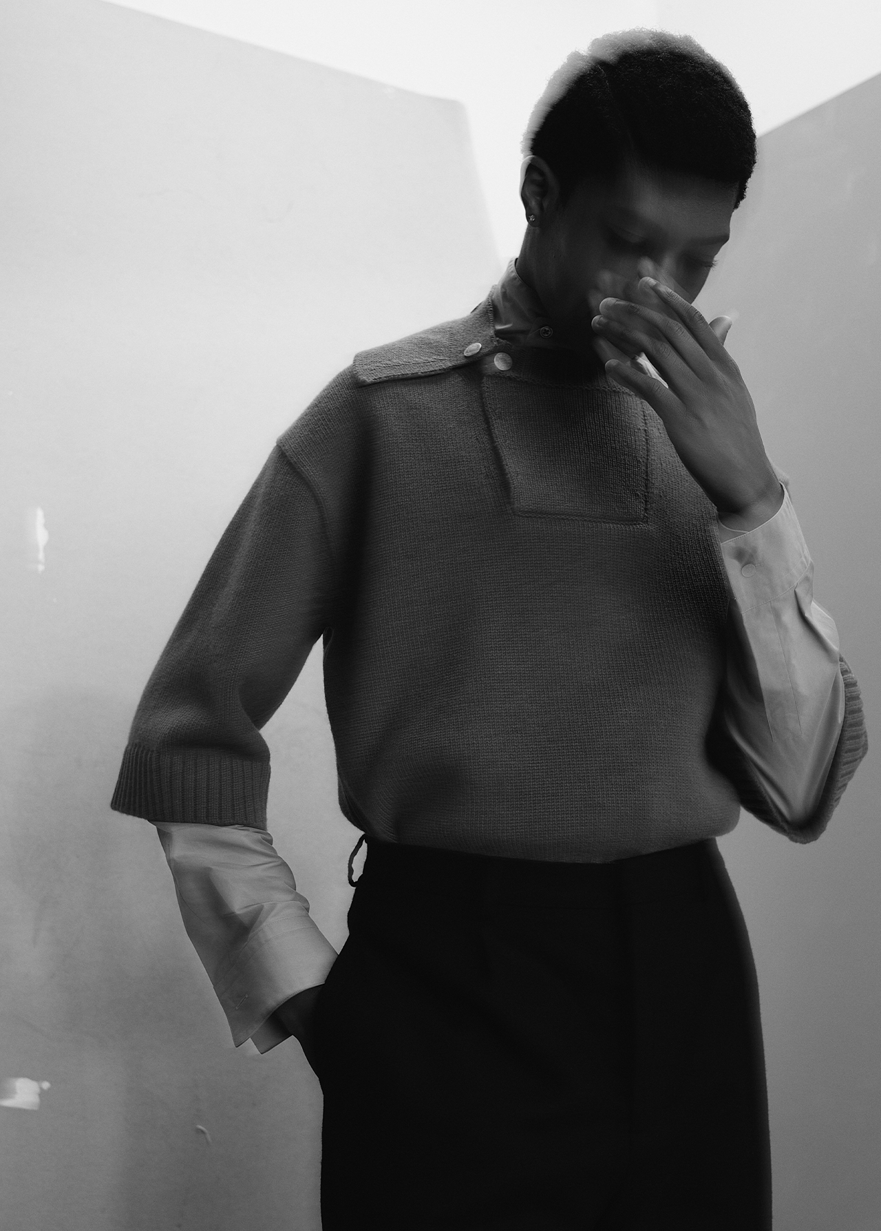 Black and white portrait of a stylish person wearing a sweater and dress pants posing against a white wall in an indoor setting.