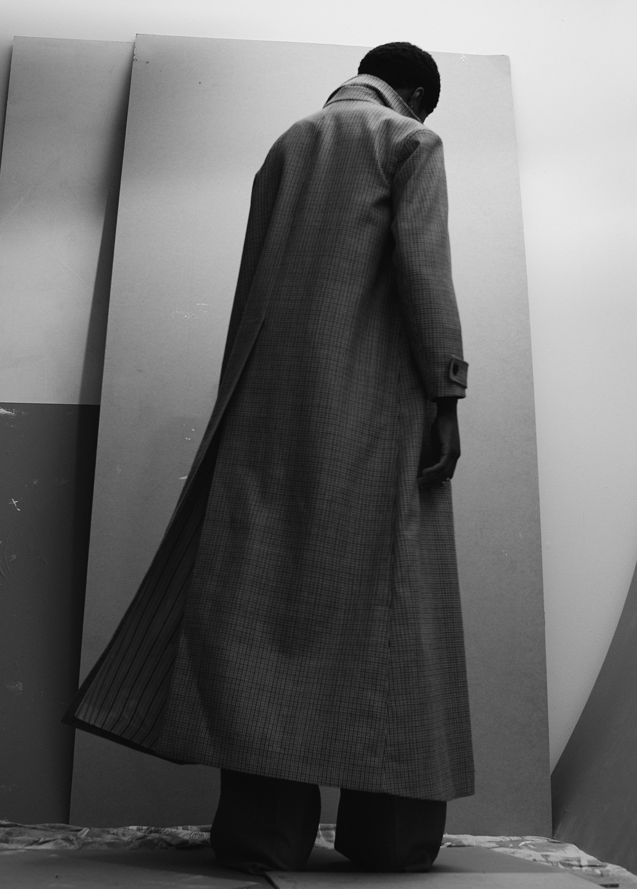Monochromatic fashion shot: a Black male model, back to camera, wears a long grey coat against a light grey backdrop. Minimalist studio.