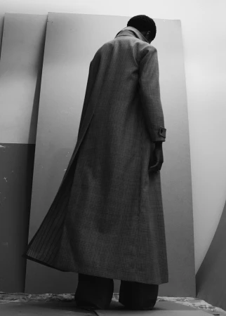Monochromatic fashion shot: a Black male model, back to camera, wears a long grey coat against a light grey backdrop. Minimalist studio.