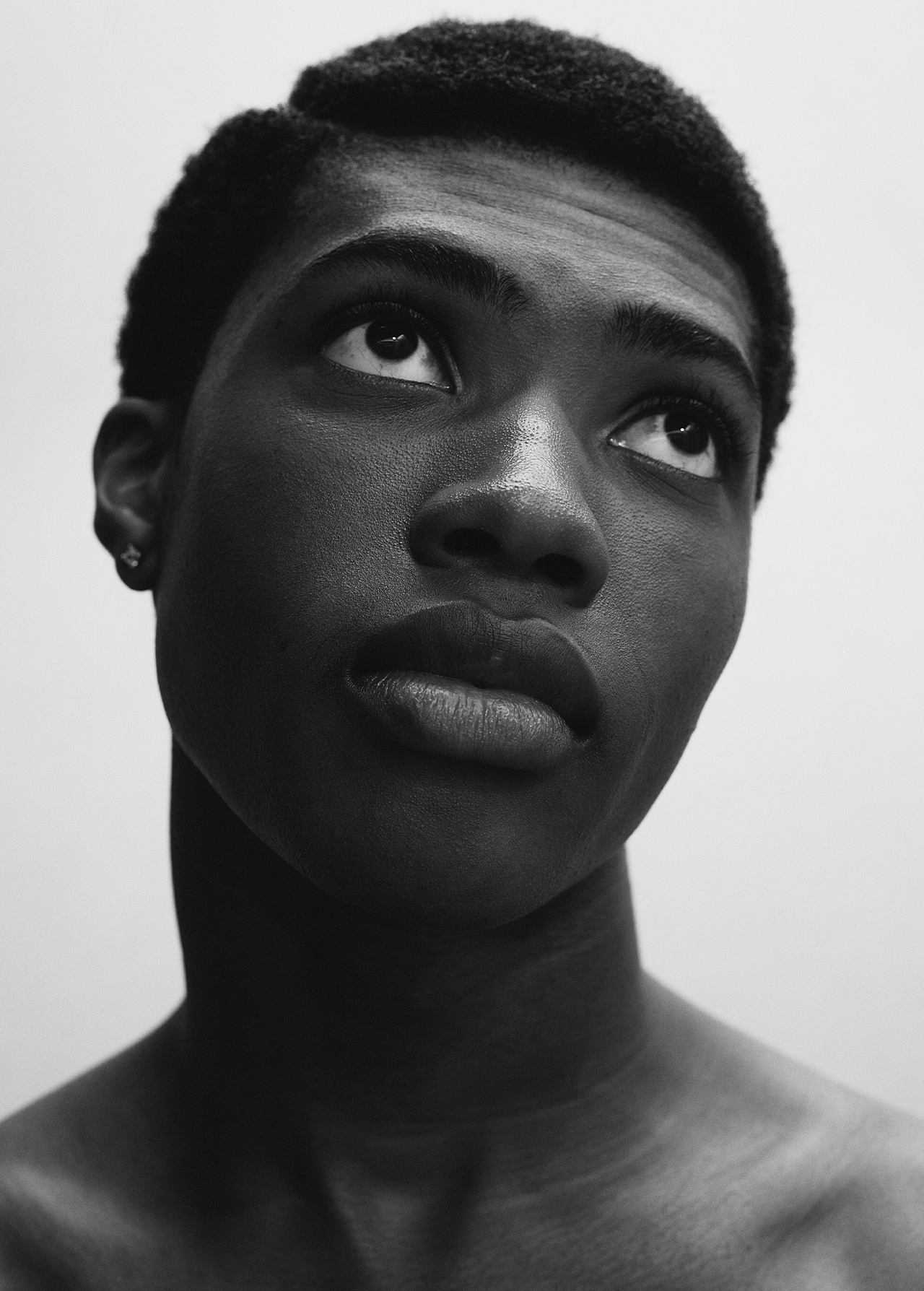 Striking portrait of a young, dark-skinned man looking upwards with a serious expression in a high-contrast black and white studio shot.