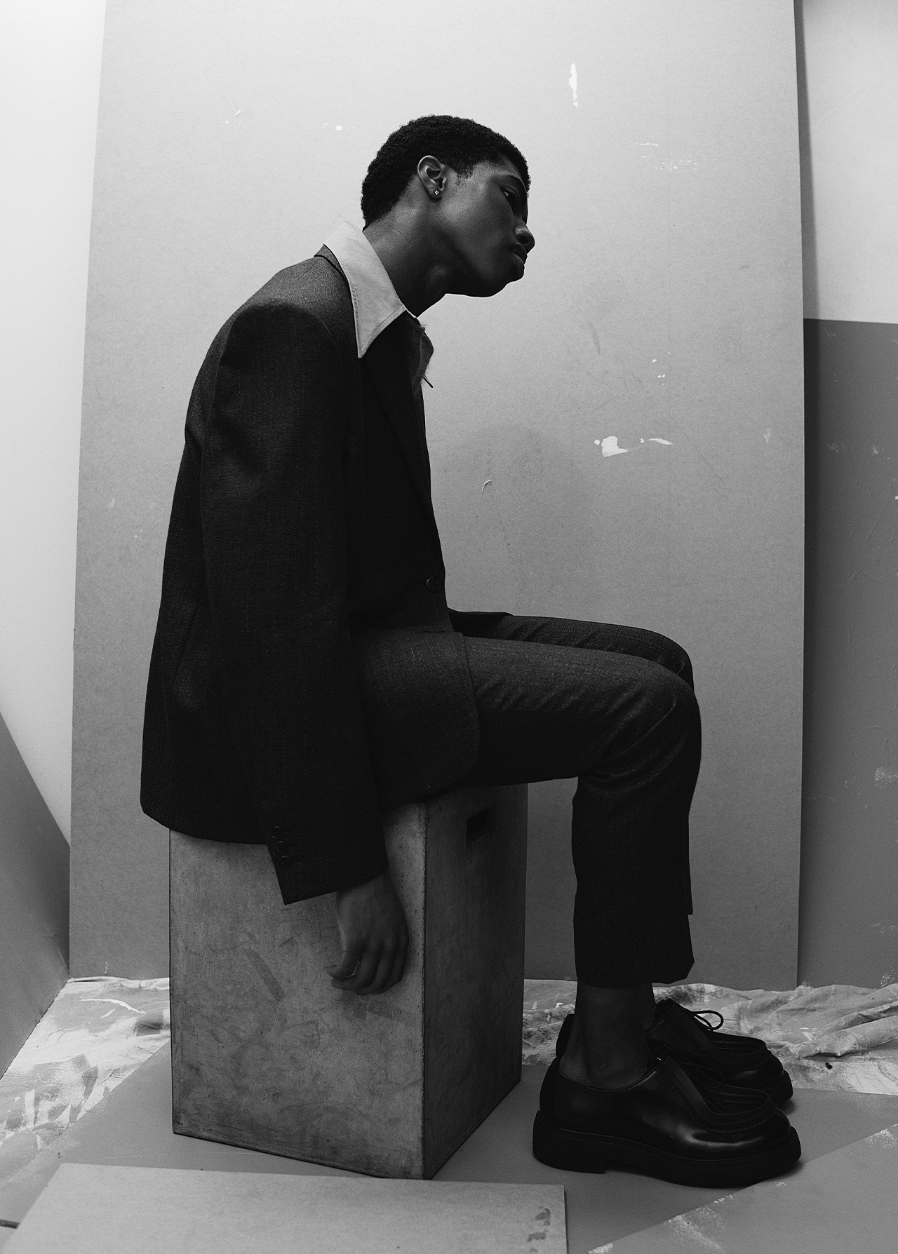 Black and white portrait of a young black man wearing a blazer, sitting on a box in a photo studio setting, looking off to the side.