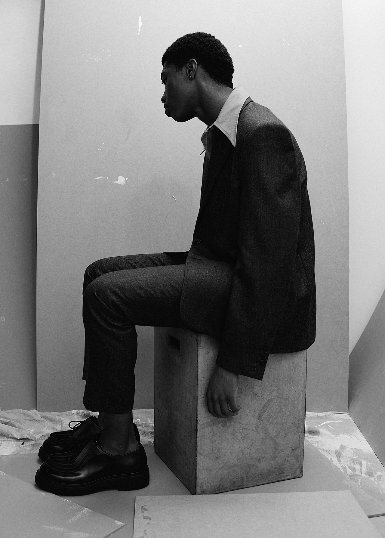 Monochromatic portrait of a young Black man in a suit sitting on a concrete block in a photo studio with neutral backdrop.