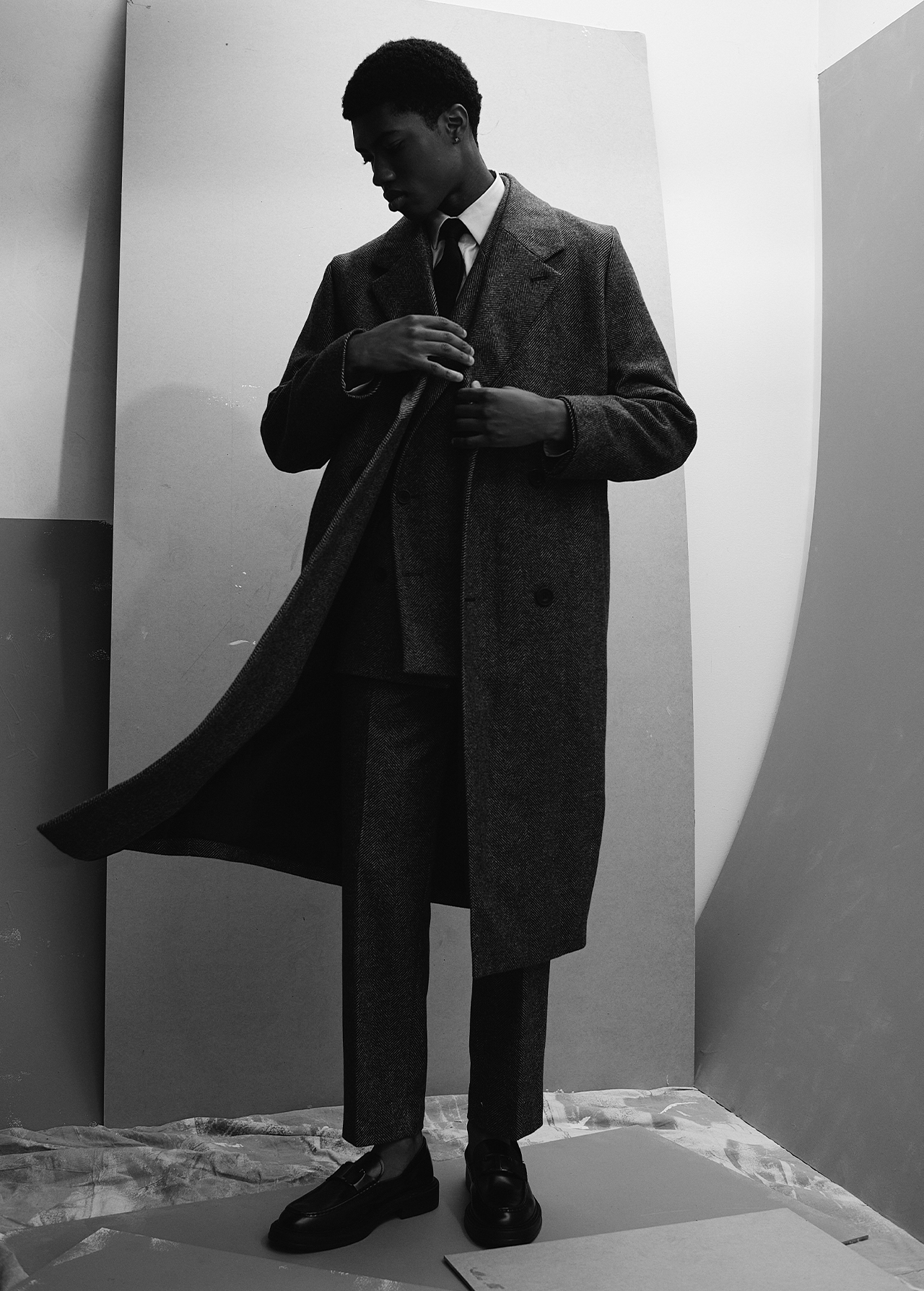 Black and white editorial portrait of a Black man wearing a tailored wool coat and suit adjusting his lapel in a studio