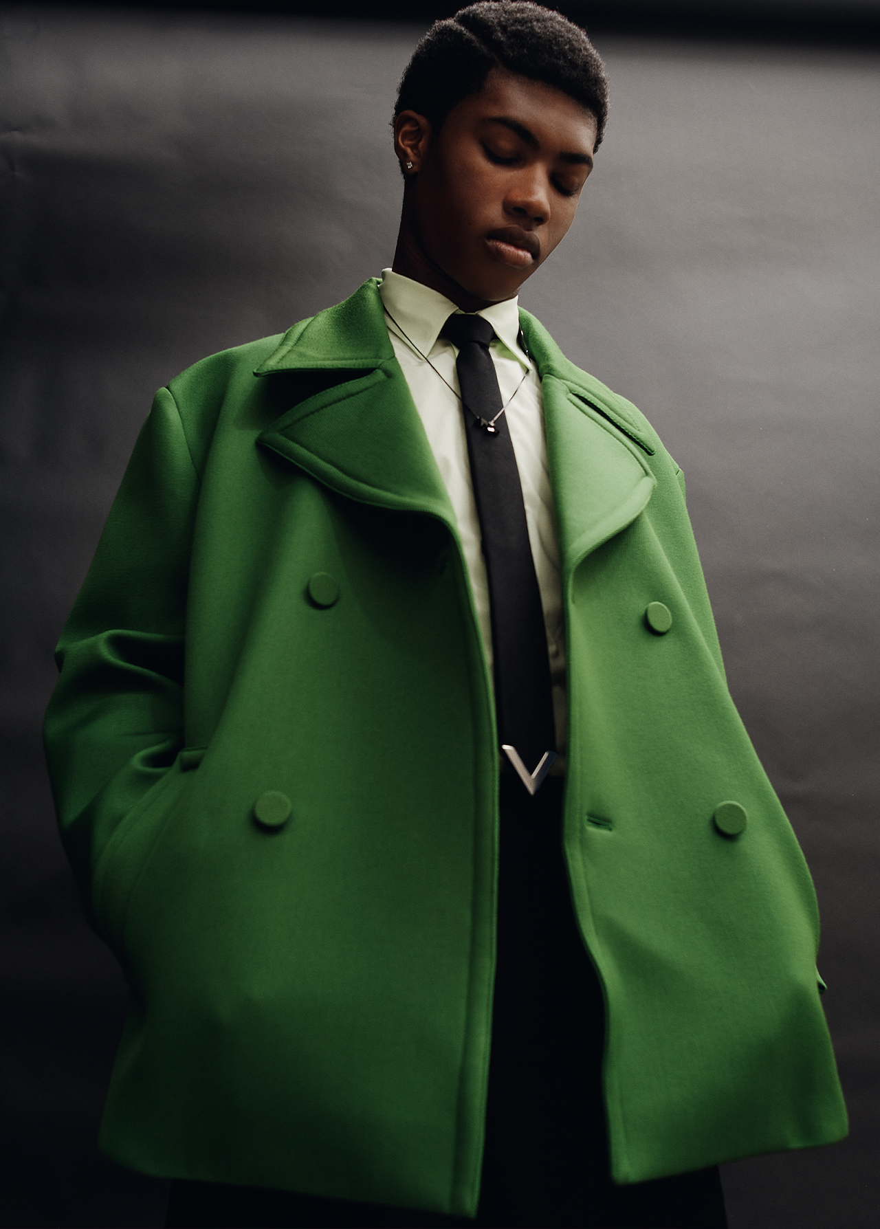 Portrait of a Black male model wearing a pea coat in vibrant green, paired with a black tie, posing against a dark backdrop.