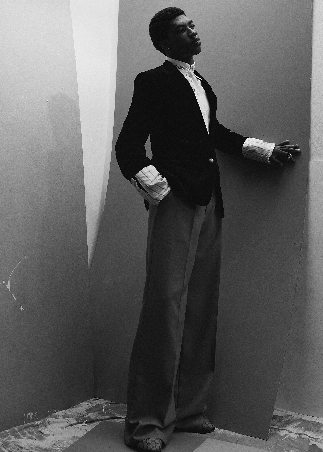 Stylish young Black male model in a velvet jacket and wide-leg pants poses in a corner photo studio with neutral-tone walls in black and white.