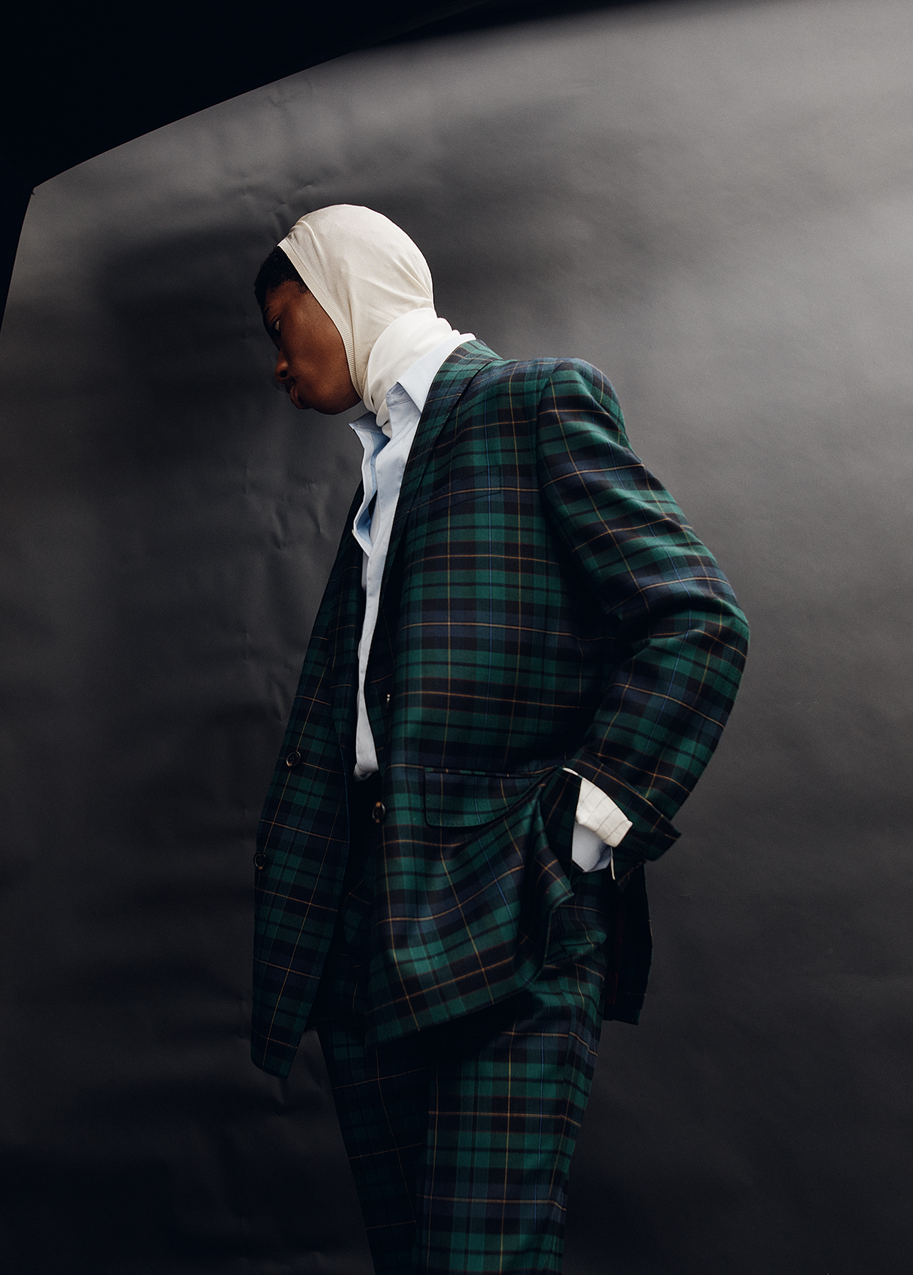 Dark-skinned model in a plaid suit with a cream-colored headscarf poses against a dark background in a studio shot.