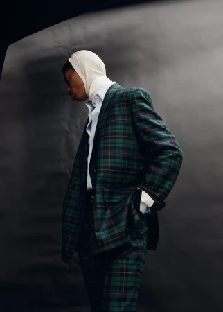 Dark-skinned model in a plaid suit with a cream-colored headscarf poses against a dark background in a studio shot.