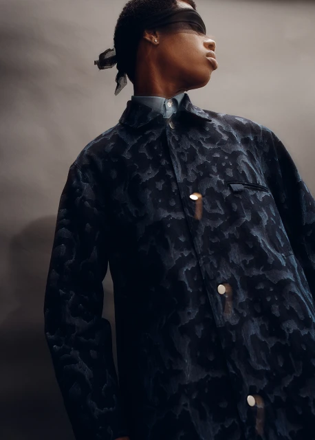 Striking fashion shot of a young, slender African American woman wearing a textured navy jacket, headscarf, and light blue shirt against a gray backdrop.
