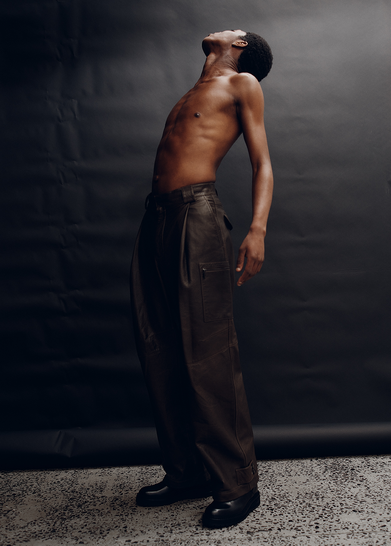 A lean, shirtless black male model poses in studio lighting while wearing dark brown pants, tilting his head back with a black backdrop.