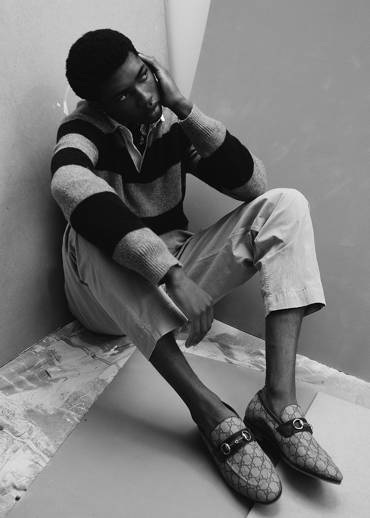 Young black male model sitting indoors, wearing striped sweater and patterned loafers, in a stylish, monochromatic fashion portrait.