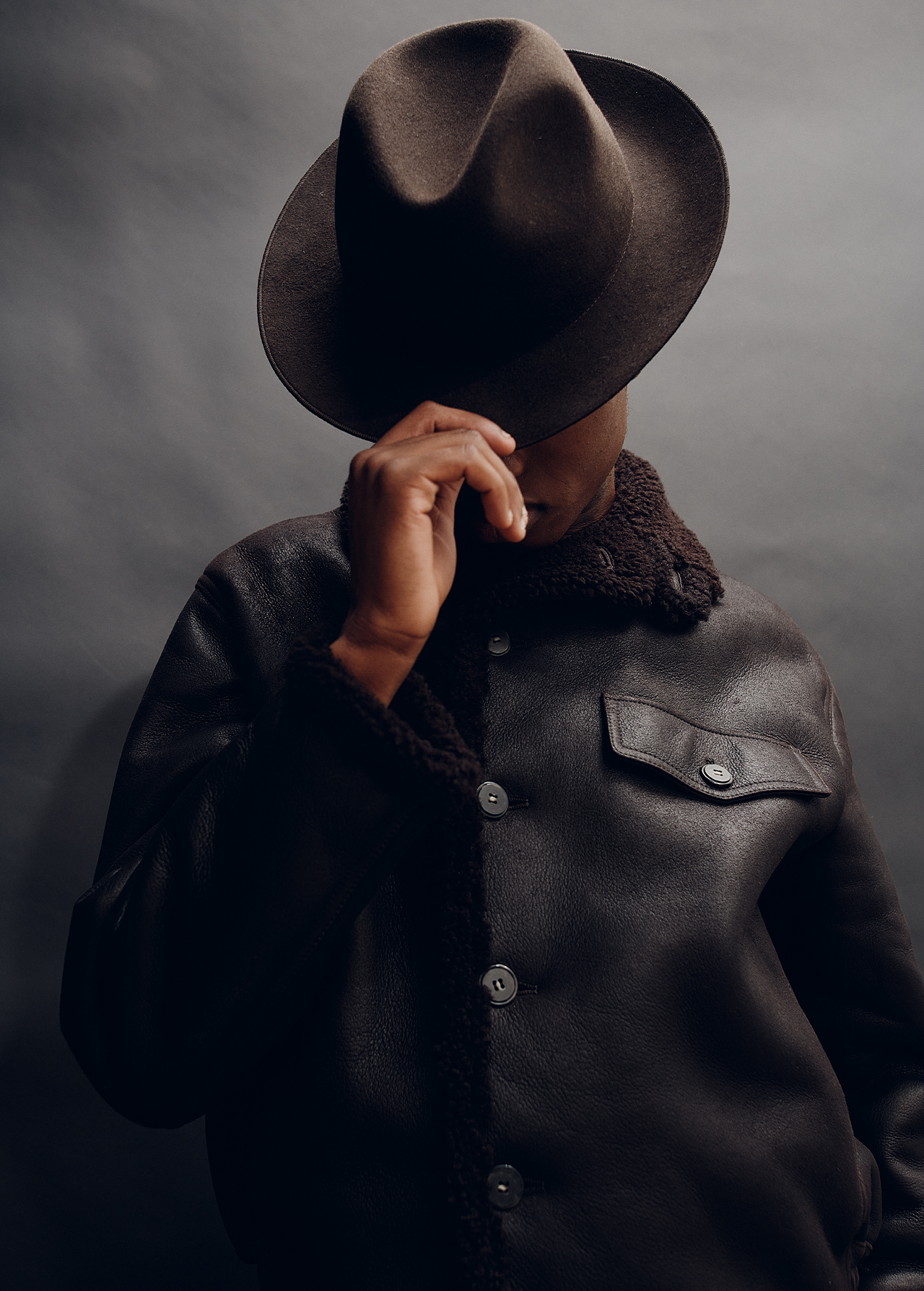 Man in shadow wearing fedora and leather jacket poses against dark background in fashion portrait. Stylish outerwear look.