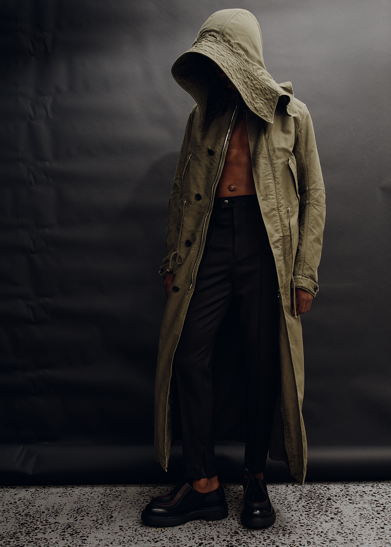 Man wearing an olive green hooded coat and dark trousers standing against a dark seamless backdrop.