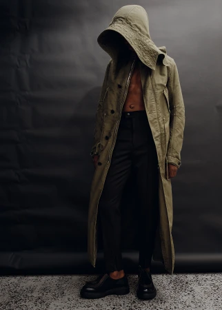 Man wearing an olive green hooded coat and dark trousers standing against a dark seamless backdrop.