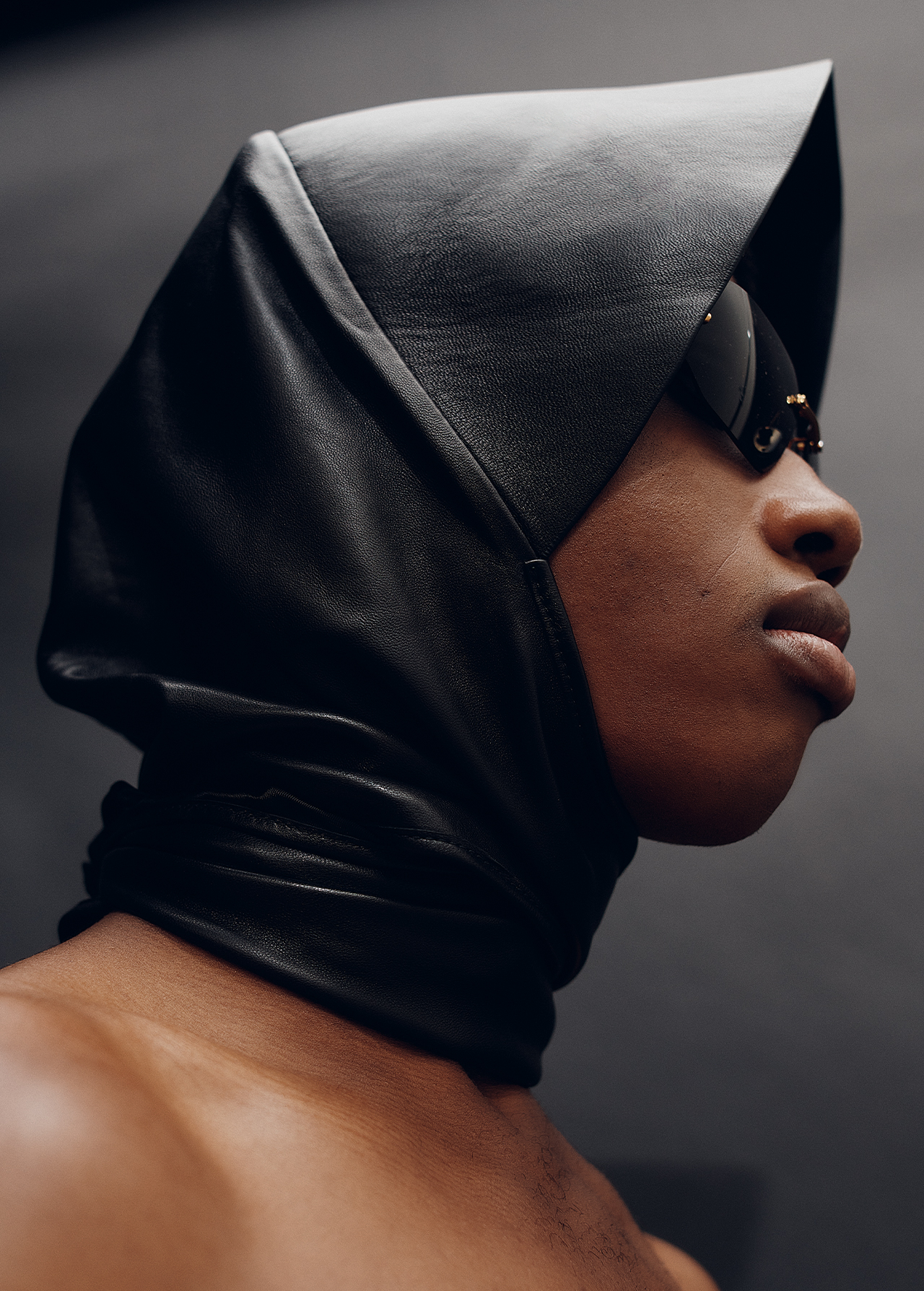 Close-up portrait of a black male wearing a black leather hood and sunglasses against a dark gray background in a studio.