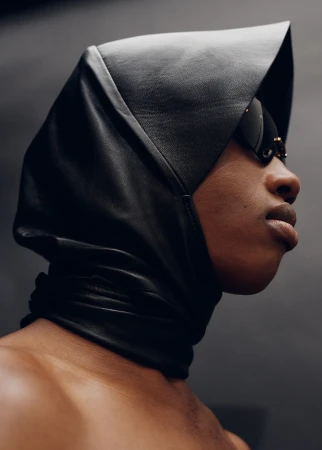 Close-up portrait of a black male wearing a black leather hood and sunglasses against a dark gray background in a studio.