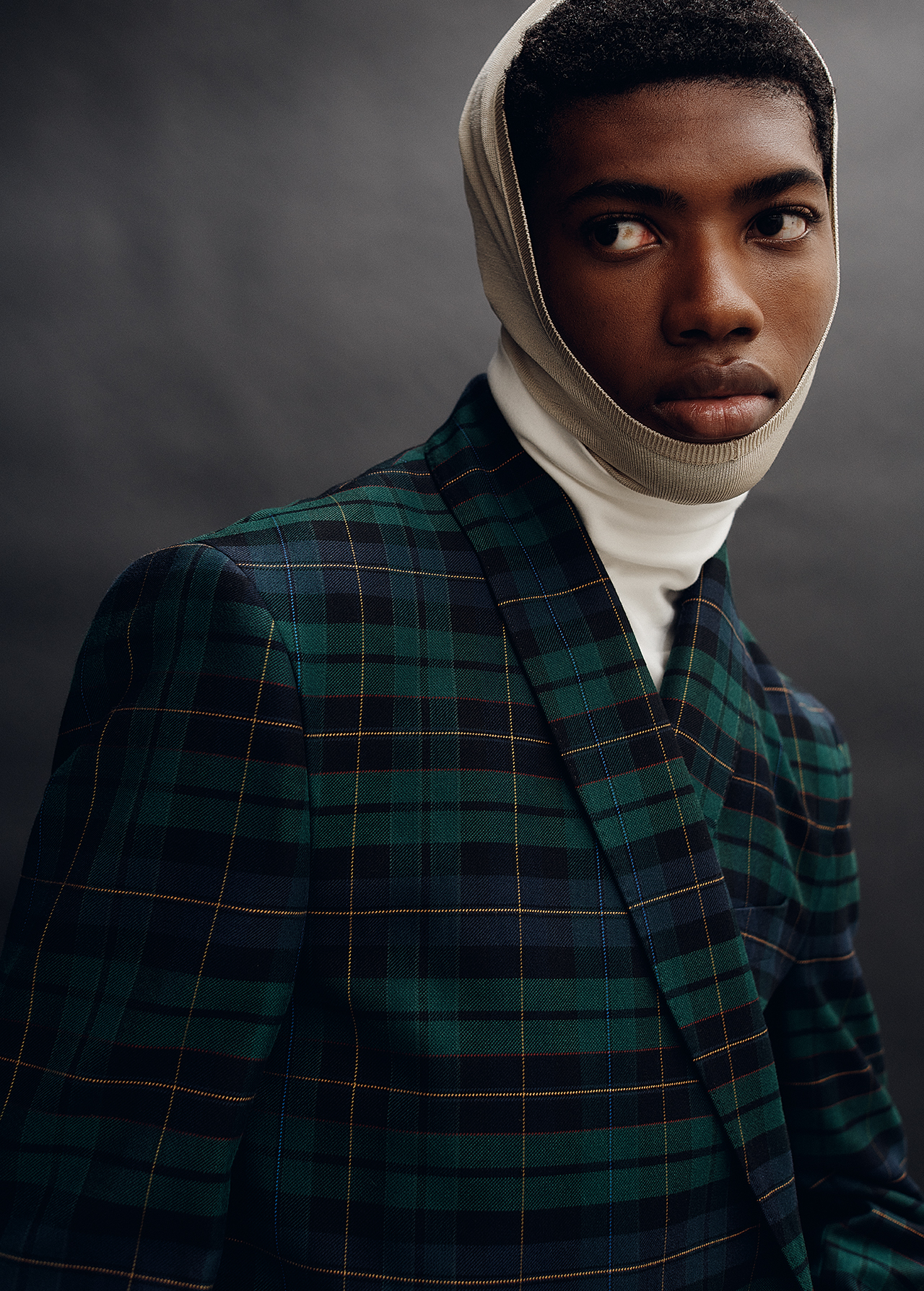 Close-up portrait of a Black man wearing a green plaid blazer, white turtleneck, and light-colored head covering against a dark background.