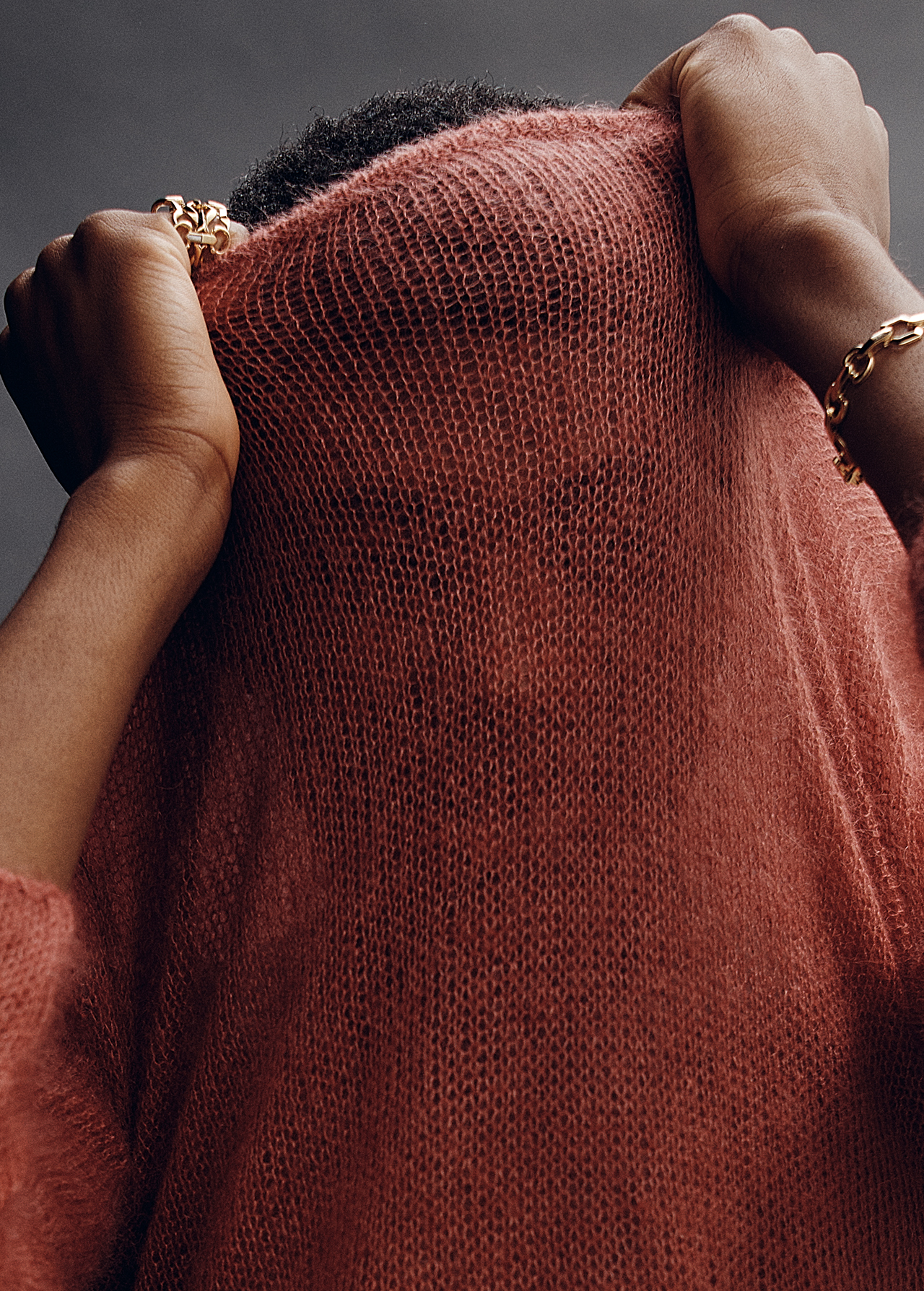 Dark-skinned person in a textured rust-colored top pulling it over their head, accessorized with gold jewelry, against a grey backdrop.
