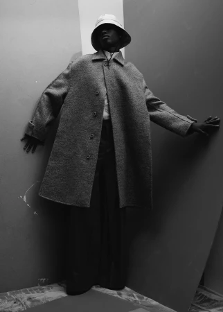 Monochrome portrait of a model in a textured long wool coat and bucket hat posing in a corner for Icon Magazine Australia.