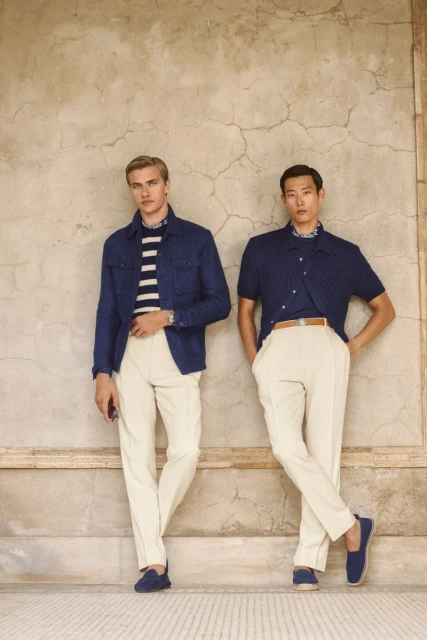 Two men pose against a textured wall, wearing stylish outfits: blue jackets, striped shirts, cream pants, and matching loafers.